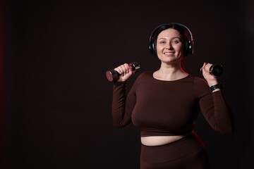 Plus size woman in gym clothes with dumbbells and headphones on black background, space for text