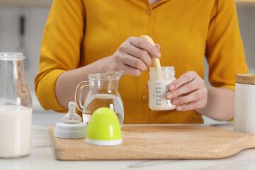 Mother making baby formula in feeding bottle at table indoors, closeup