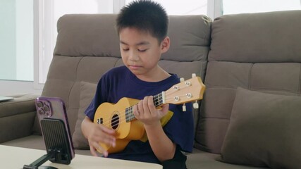 A young boy sits on a couch playing a ukulele, streaming an online music lesson via a smartphone mounted on a stand. The cozy home setting highlights interactive learning and creative expression. - Powered by Adobe