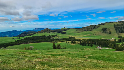 Obraz premium Verdant hills roll across Plankogel, Styria, Austria, under a partly cloudy sky. Patches of coniferous trees dot the landscape, creating a textured, scenic view of the Austrian countryside. Tranquil