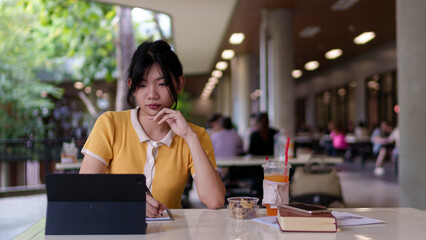 Young Asian woman studying with a tablet and book in a university cafe