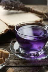 Aromatic lavender tea in glass cup, brown sugar, book and dry flowers on wooden table, closeup