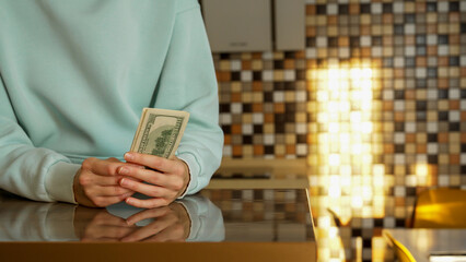 Light streams into a kitchen as a person in a light blue sweatshirt holds a rolled bundle of cash in their hands, highlighting the modern interior and colorful tile walls.