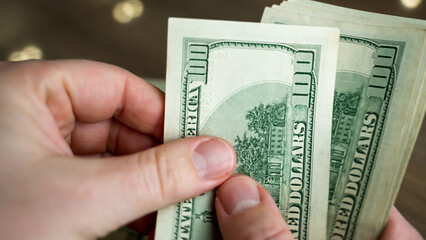 Hands with light skin are counting several hundred dollar bills in a well-lit indoor space. The bills are held firmly while focusing on their details and texture.