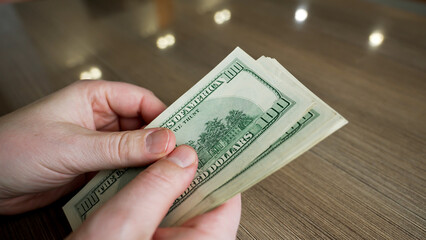 A pair of hands is seen counting several one hundred dollar bills at a wooden table. The setting is bright with natural light coming through nearby windows.
