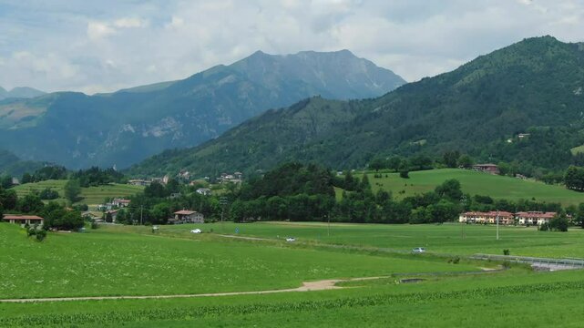 Busy road crossing Clusone plateau, green fields and meadows with mountains in background, Rovetta, Italy. Aerial forward ascending