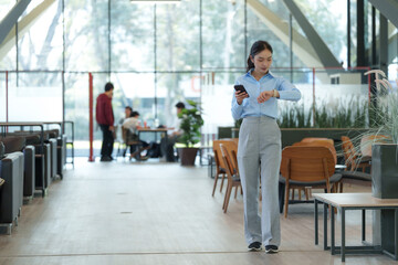 Young asian businesswoman walking in modern office hallway checking the time on her smartwatch while using her smartphone, with colleagues working in the background