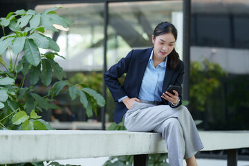 Obraz premium Young asian businesswoman is using her smartphone while sitting on a bench in the atrium of a modern office building, taking a break from work and enjoying the greenery