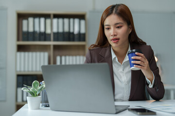 Young asian businesswoman sipping coffee while working on a laptop in a modern office, highlighting...