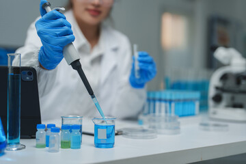 Scientist wearing lab coat and gloves using micropipette dropping blue liquid in beaker during...
