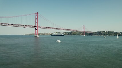 The ship sails under a red suspension bridge on a wide river in Portugal. Yachts and sailboats sail on the river. Drone video.