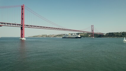 The ship sails under a red suspension bridge on a wide river in Portugal. Yachts and sailboats sail...