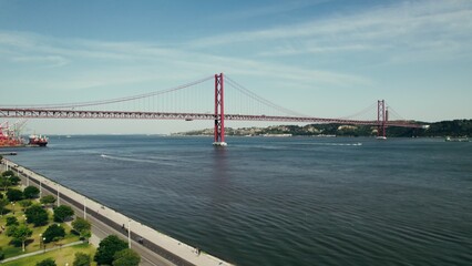Panoramic bird's-eye view of a wide river with a red suspension bridge connecting the banks. The drone is flying over the embankment