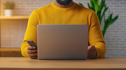 Man in yellow sweater using laptop and smartphone at desk