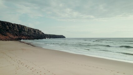 The drone flies over the sea, the sea waves wash the sandy beach lying at the foot of a rocky mountain