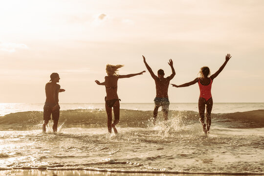Group of four happy young friends runs to sunset sea beach and jumps over waves with splashes. Tropical vacations concept