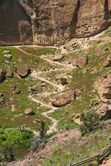 A winding path through rocky terrain leads hikers to breathtaking views, surrounded by unique rock formations and lush greenery under a bright sunny sky.