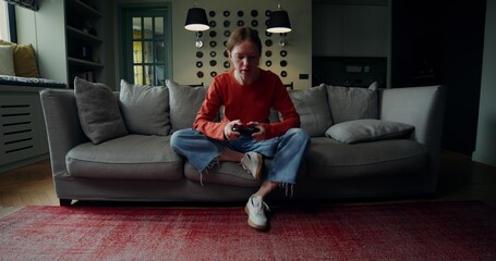 Obraz premium A young woman in casual clothes is smiling while playing video games with a joystick, sitting on the couch at home. The video moves from the general plan to the woman's face