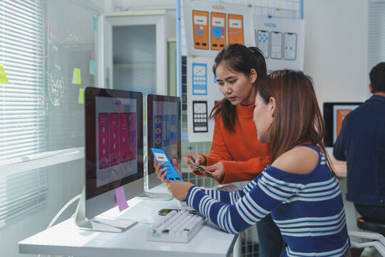 Two female web designers are comparing a mobile phone screen with the design displayed on a computer screen, collaborating on user interface and user experience development for a mobile app