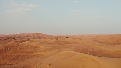 Drone view of a lifeless desert with sparse vegetation among red sand. Cars drive into a sandy mountain in the background
