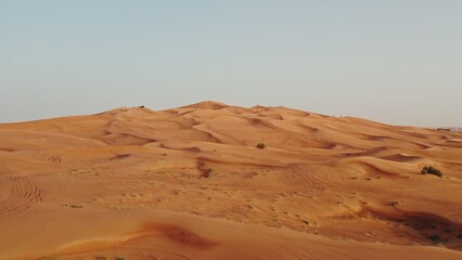 Drone view of a lifeless desert with sparse vegetation among red sand. Cars are standing on a sandy mountain in the background