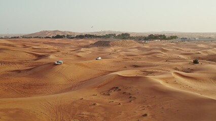 Cars drive off-road on the red sand dunes of the desert with sparse vegetation among the sand, drone video