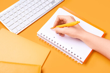 Female hand with pencil writing in notepad on orange background