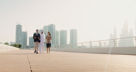 A man in traditional white Arab clothes with a laptop in his hands communicates with European business partners, walking down the street of a modern city