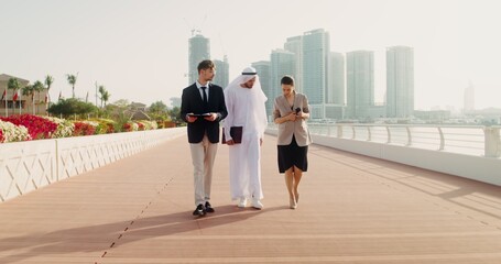 A man in traditional white Arab clothes with a laptop in his hands communicates with European business partners, walking down the street of a modern city
