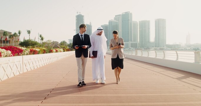 A man in traditional white Arab clothes with a laptop in his hands communicates with European business partners, walking down the street of a modern city