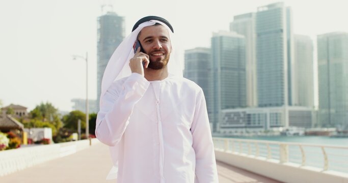A young man in Arab national white clothes smiles, while talking on the phone walking along the embankment of a modern city on a sunny day