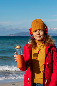 young woman with headphones drinking water by the sea in cold weather