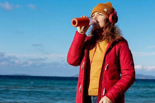 young woman with headphones drinking water by the sea in cold weather