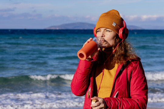 young woman with headphones drinking water by the sea in cold weather
