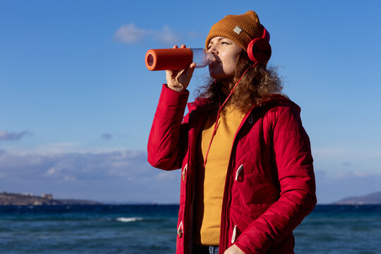 Cute woman with headphones drinking water by the sea in cold weather