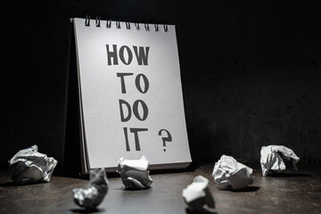 oświetlony otwarty brulion ze słowem HOW TO DO IT oraz znakiem zapytania, na ciemnym tle, dookoła rozrzucone zmięte kartki papieru © siwyk