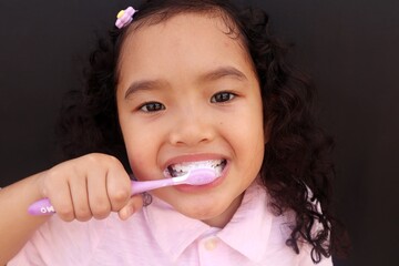 Asian toddler girl brushing her own teeth, close-up view on dark background.