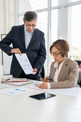 Senior professionals work on project in modern office, analyzing documents and charts. The man focuses intently the woman reviews papers with calculator ,dynamic scene of collaboration and expertise.