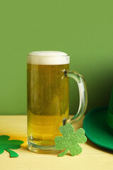 Mug of beer with leprechaun hat and paper clovers on yellow wooden table against green background....