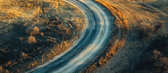 Aerial view of a winding rustic road with earth tones and a textured surface surrounded by dry grass and sparse shrubs under soft evening light
