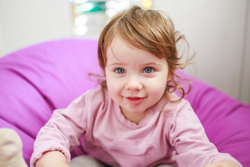 Fototapeta premium A young girl with light brown hair and blue eyes is smiling. She is seated on a purple bean bag chair, creating a playful atmosphere. The background is softly out of focus