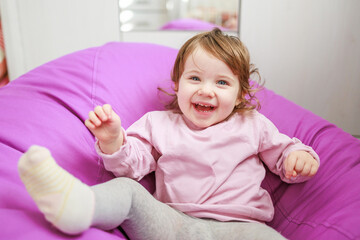 Fototapeta premium A young girl with light brown hair and blue eyes is smiling. She is seated on a purple bean bag chair, creating a playful atmosphere. The background is softly out of focus