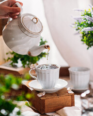 A hand pours tea from a white porcelain teapot into a matching white cup, set on a saucer. The...