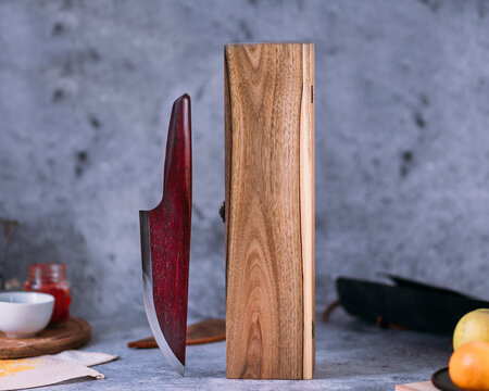 A knife with a polished, dark reddish-brown wooden handle and a sleek metal blade rests on a textured gray surface. Nearby, there's a brown leather sheath and a wooden box