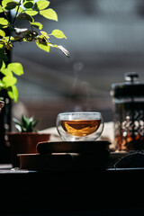 A transparent cup filled with tea sits on a windowsill, with visible steam rising above it. The cup...