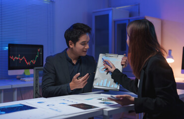 Two asian business partners reviewing financial charts and graphs on a clipboard, discussing investment strategies and analyzing market trends in a brightly lit office late at night