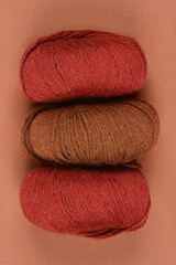 A top-down shot of three balls of yarn in varying shades of brown and red, arranged in a row against a solid brown background, showcasing their texture and color tones