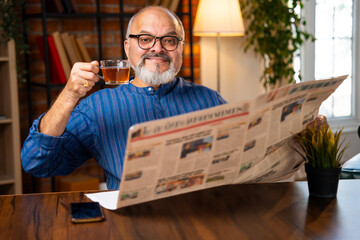 Retired Indian senior man in kurta reads daily newspaper at home desk with black coffee