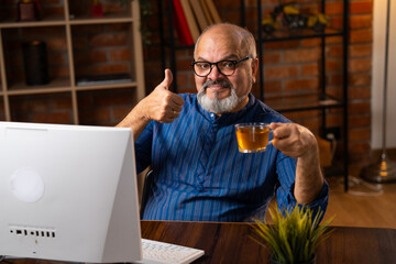Retired Indian senior man in kurta uses desktop computer to type work reports at home workstation