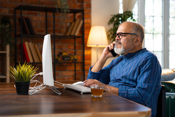 Indian elderly man in kurta works on desktop, talks on the phone, and drinks coffee at home